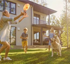 A sunny outdoor scene showing a woman and children playing with a frisbee, while a dog runs through the grass near a house.