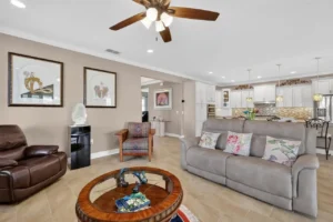 A cozy living room with two sofas, a round coffee table, wall art, and a ceiling fan, leading to a modern kitchen.