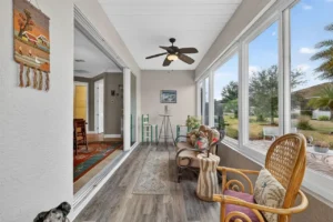 A light and airy sunroom with large windows, casual seating, wooden flooring, and a ceiling fan, surrounded by greenery.