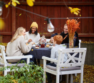 A group of four people enjoying a meal outdoors, surrounded by autumn leaves and a cozy setting with decorative plants and a jar of drink.