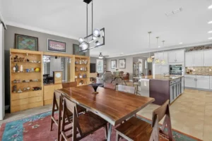 A modern dining area featuring a large wooden table, stylish lighting, and an open layout connecting to a kitchen with light cabinets.