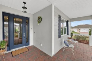 A welcoming front porch with a decorative wreath, potted plants, a doormat, and seating, framed by blue doors and light gray walls.