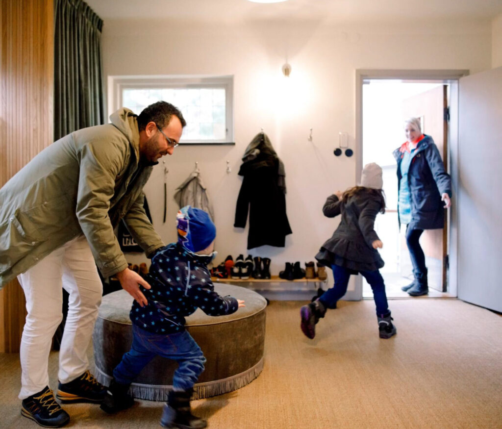 A man playfully interacts with a child in a hallway, while another child runs towards an open door where an adult waits.